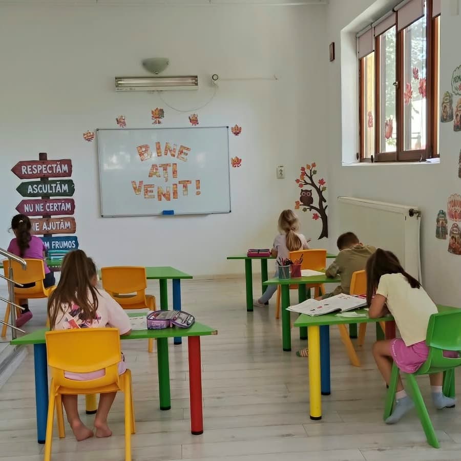 Copii la mese, făcând teme, în sala de after school decorată cu tabla de bun venit