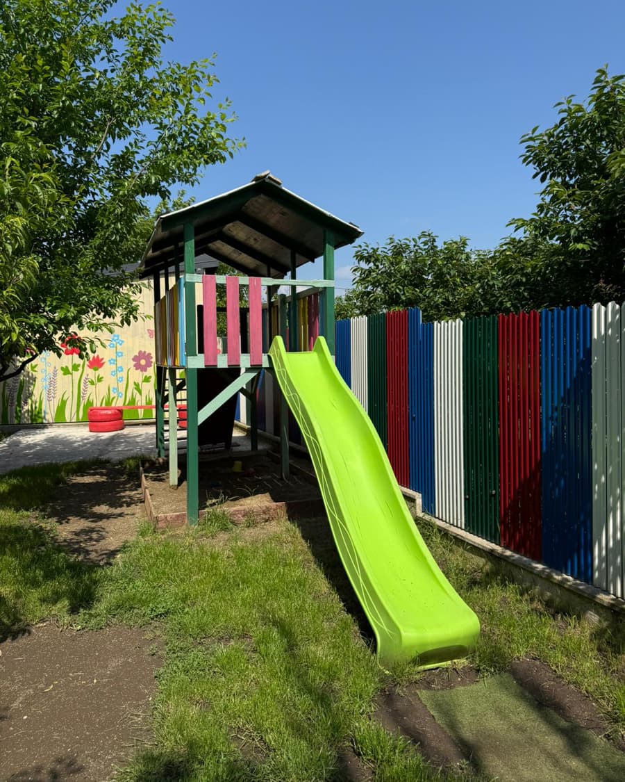 Turn de joacă verde cu tobogan, lângă gardul viu colorat al curții