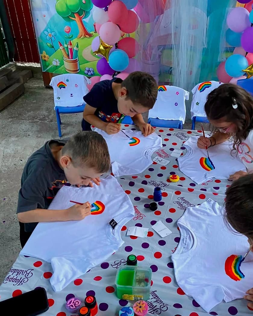 Copii pictând tricouri albe la o masă lungă acoperită cu folie