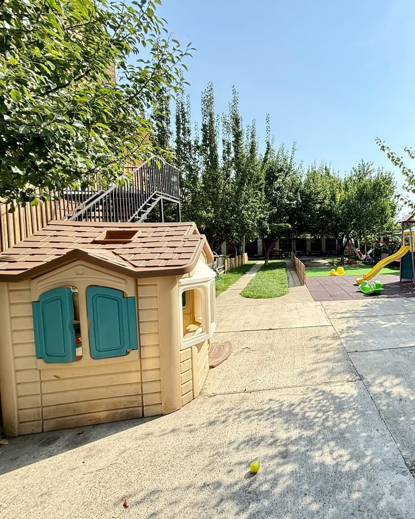 Căsuța de joacă, cu obloane turcoaz, lângă locul de joacă cu tobogan și leagăne